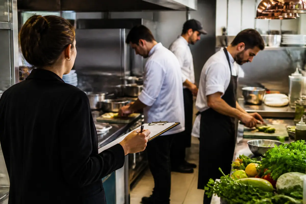Inspectrice réalisant un contrôle d'hygiène restaurant en cuisine avec chefs cuisinant aliments frais